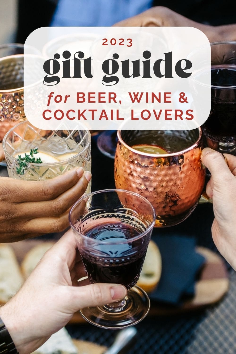 Side view, hands holding wine glasses and cocktail glasses coming together to click glasses over a table; text overlay reads "2023 gift guide for beer, wine, and cocktail lovers".