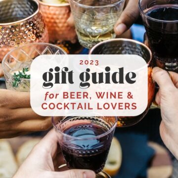 Side view, hands holding wine glasses and cocktail glasses coming together to click glasses over a table; text overlay reads "2023 gift guide for beer, wine, and cocktail lovers".