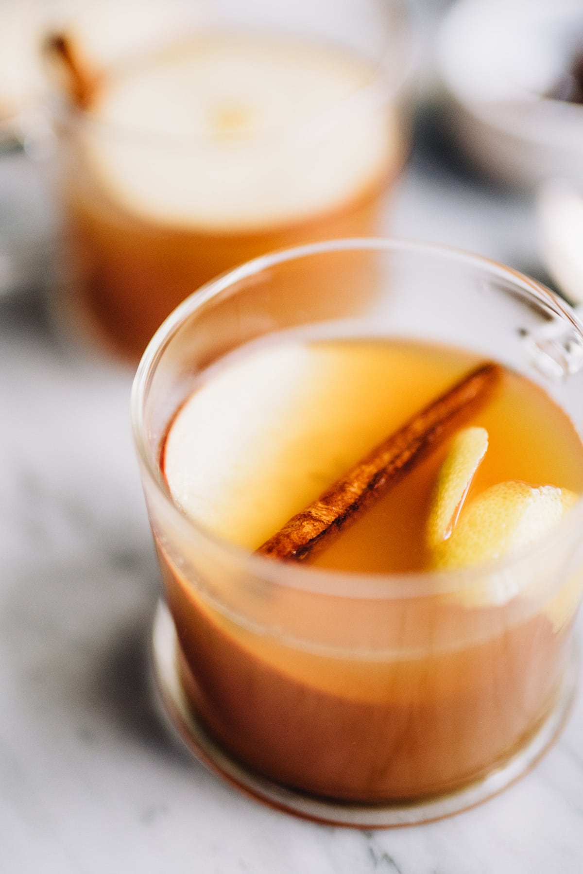Side view, two apple cider hot toddy cocktails in glass mugs, garnished with apple slices and a cinnamon stick.