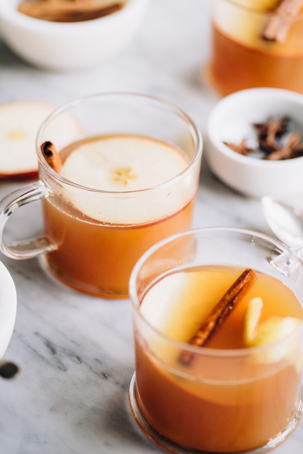 Side view, several apple cider hot toddy drinks on a marble table surrounded by small bowls filled with apple slices, cinnamon sticks, and star anise.
