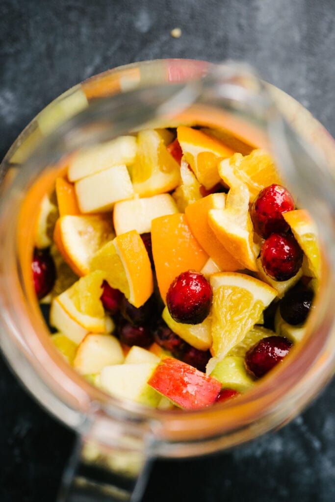 From overhead, diced oranges, apples, and fresh cranberries tossed with sugar in a glass pitcher.