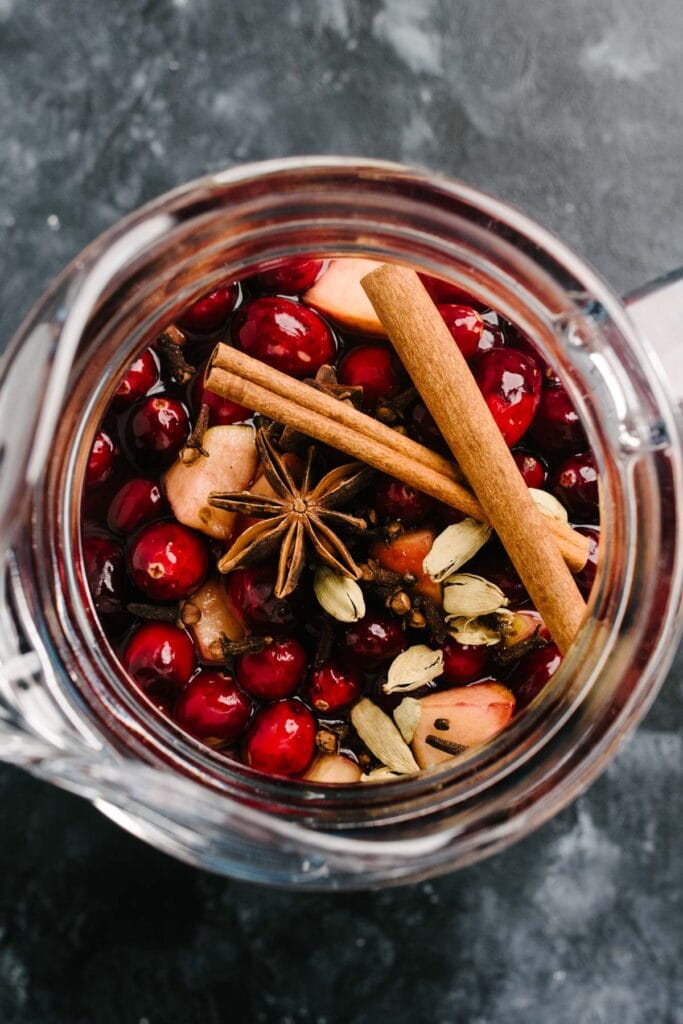 From overhead, fresh cranberries, diced apples, diced oranges, and whole spices (star anise, cinnamon sticks, cloves, and cardamom pods) in a glass pitcher.