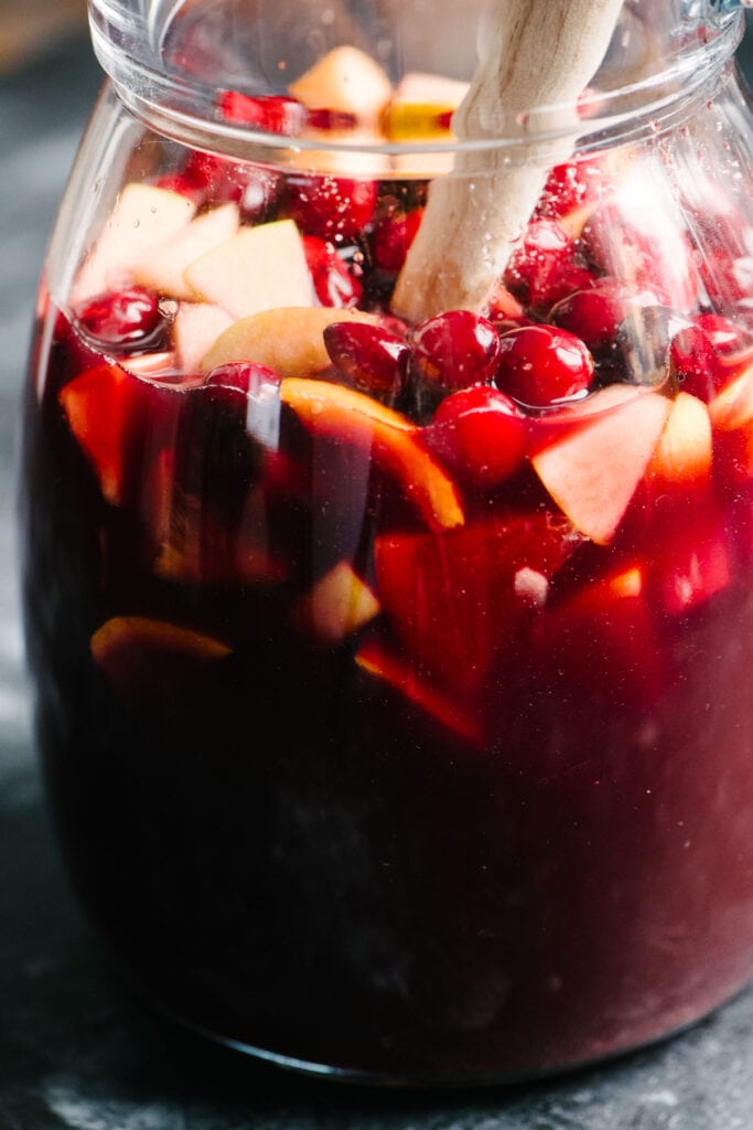 Side view, red wine holiday sangria in a glass pitcher being stirred with a wood spoon.