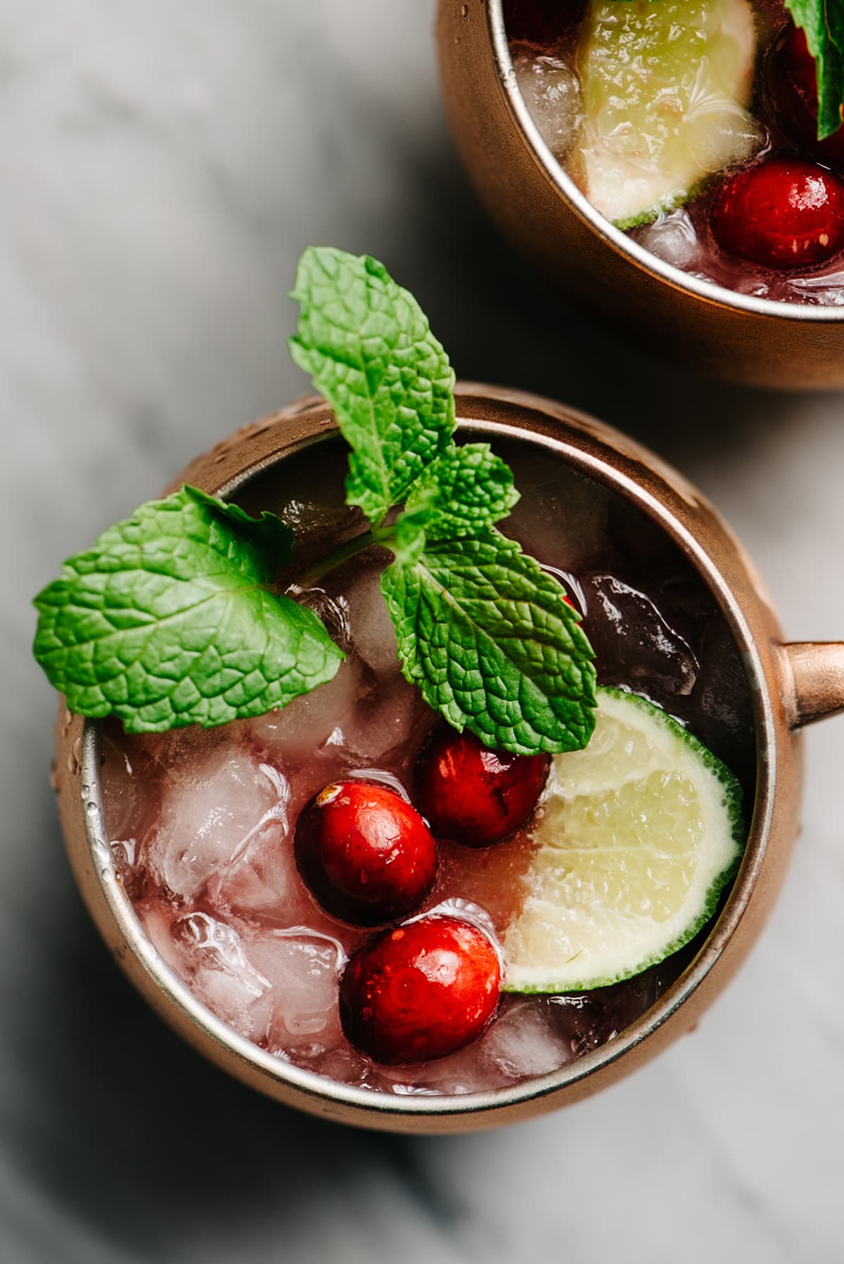 From overhead, two cranberry Moscow mule cocktails in copper mugs on a marble table, garnished with a lime wedge, mint sprig, and fresh cranberries.