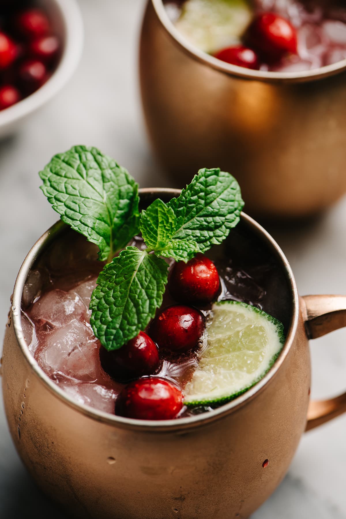 Side view, a Moscow mule with homemade cranberry syrup in a copper mug, garnished with a lime wedge, mint sprig, and fresh cranberries; a second cocktail and a small bowl of cranberries are in the background.