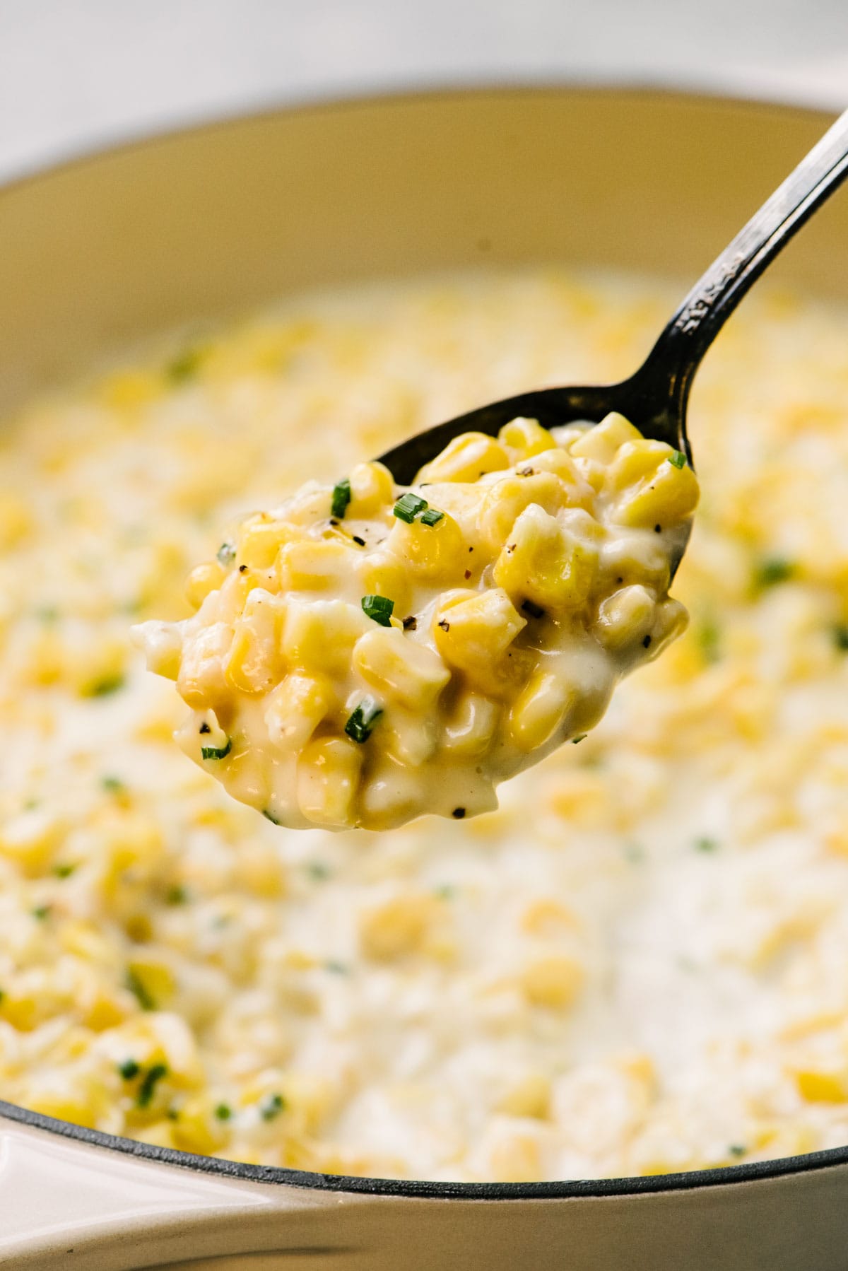 Side view, a serving spoon loaded with creamed corn hovering over a skillet.