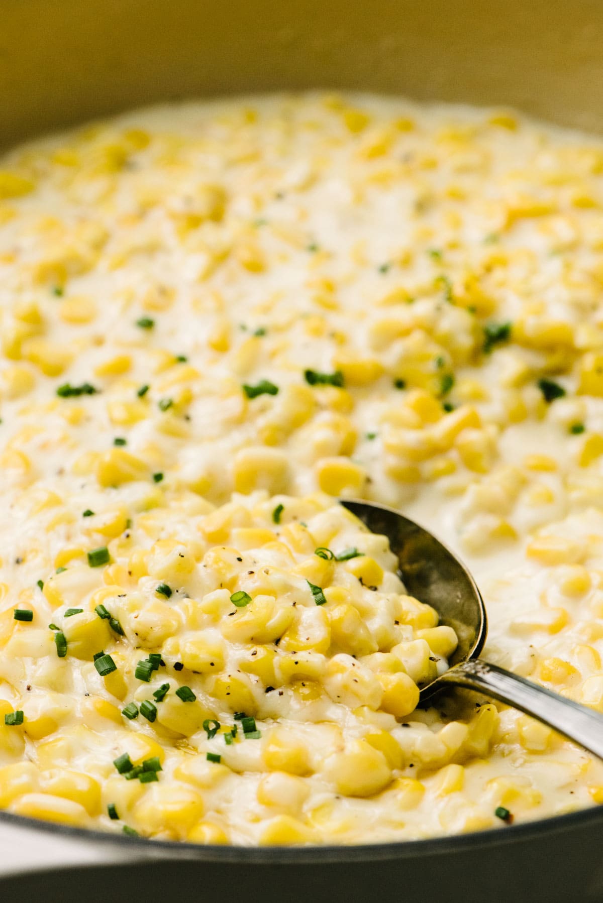 Side view, a serving spoon tucked into a skillet of homemade cream style corn, garnished with fresh chives.