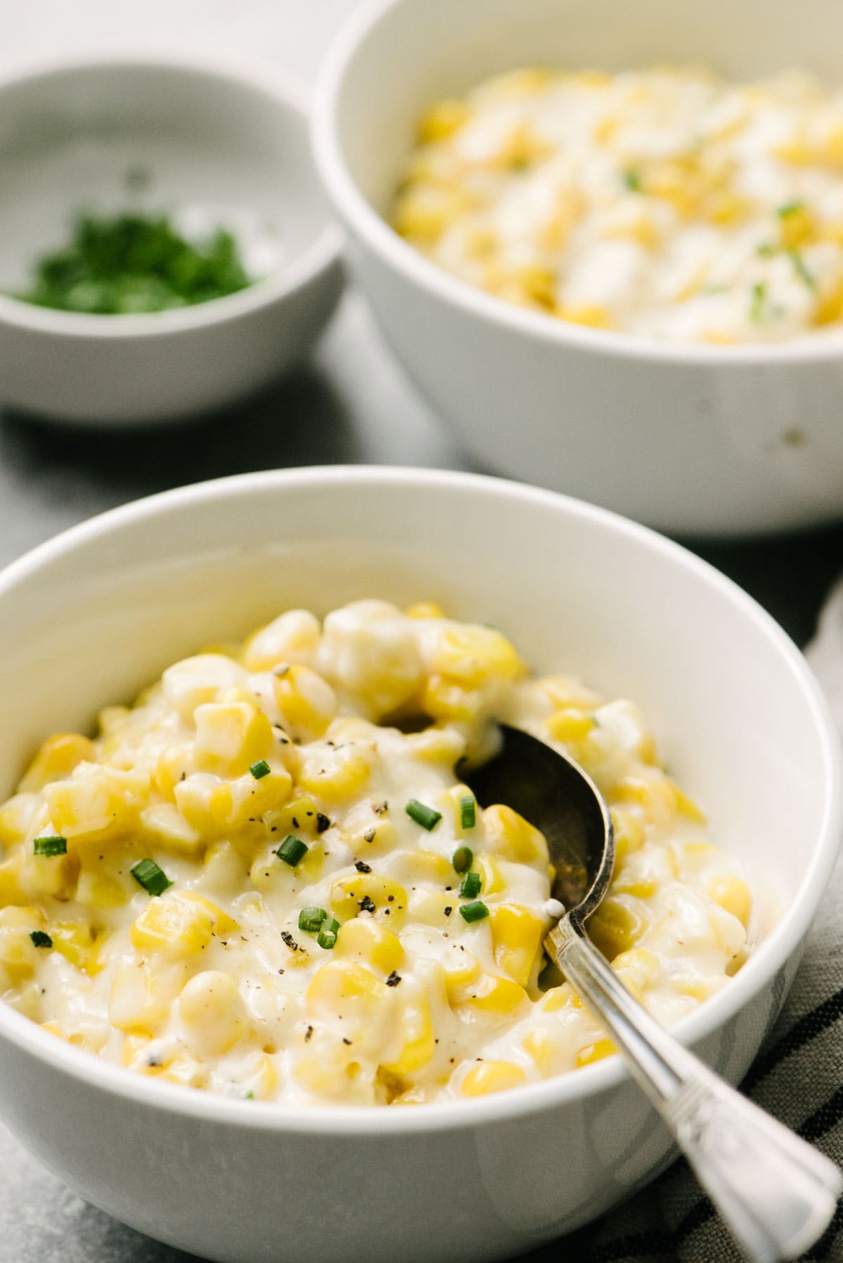Side view, a spoon tucked into a small bowl of creamed corn, garnished with fresh chives. A separate bowl and a small pinch bowl of fresh chives are in the background.