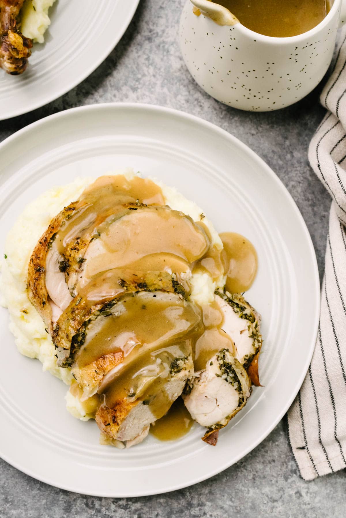 Herb roasted chicken breast slices over mashed potatoes, smothered with gravy; a striped linen napkin and small pitcher of gravy surround the plate.