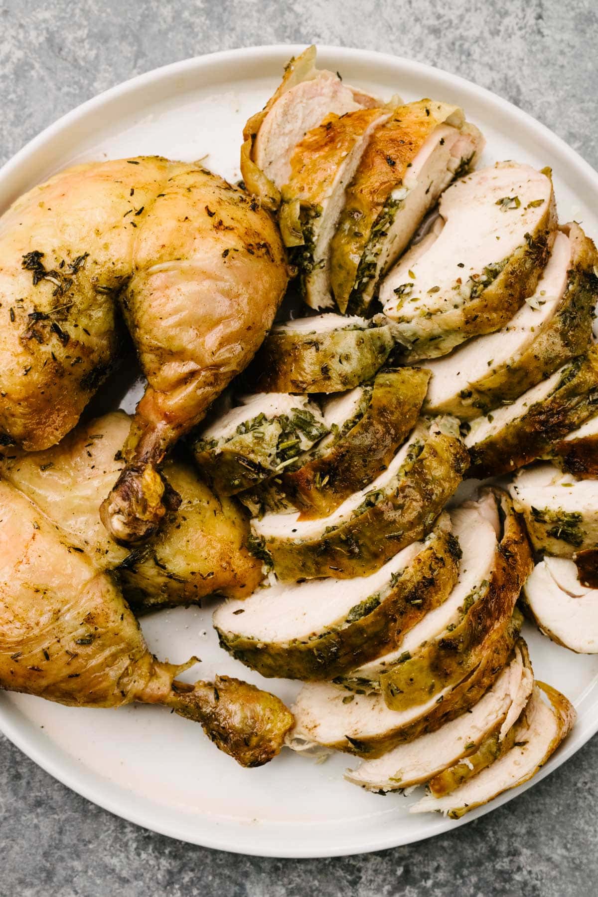 A carved herb roasted chicken on a white plate - two chicken thighs with legs attached and sliced pieces of chicken breast.