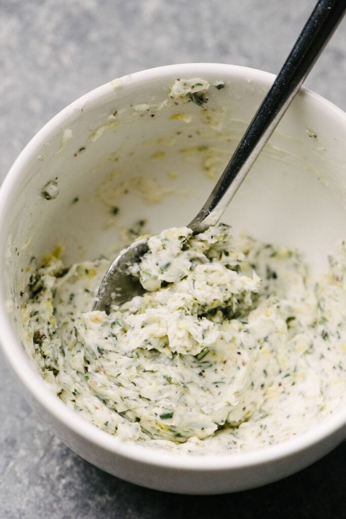 A spoon tucked into a white bowl of herb butter made from softened butter, garlic, salt, and fresh herbs.