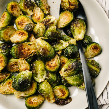 A black serving spoon tucked into roasted Brussels sprouts in a low tan serving bowl, with a striped linen tucked to the side.