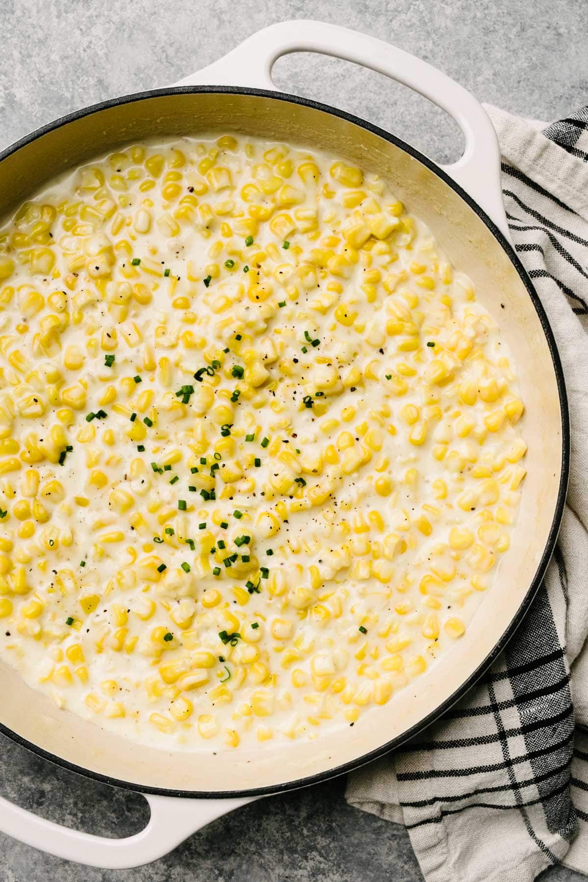 Homemade creamed corn in a large grey enameled skillet, garnished with fresh chives; a striped tan and black linen napkin is tucked to the side.