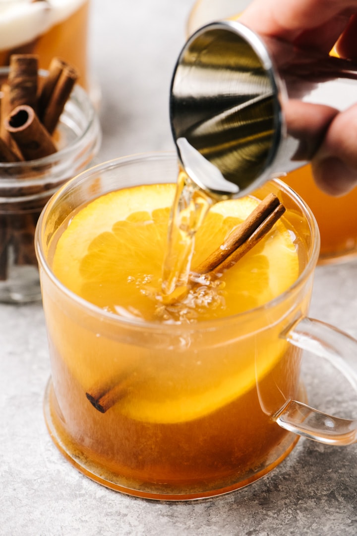 Side view, pouring rum into a glass mug of mulled apple cider with an orange slice and a cinnamon stick.