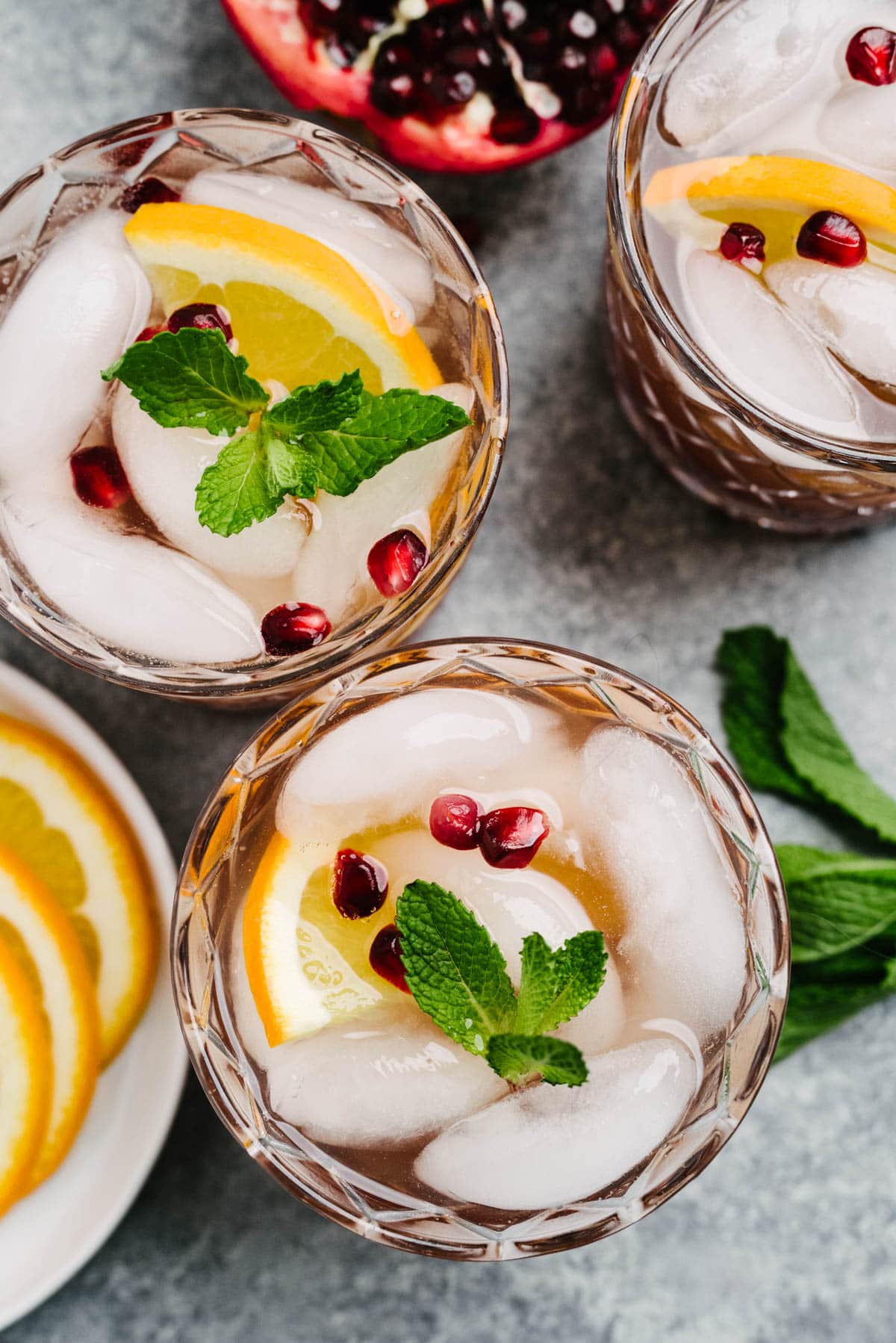 From overhead - three glasses of Prosecco punch on a concrete background, surrounded by a halved pomegranate, mint sprig, and orange slices on a white plate.