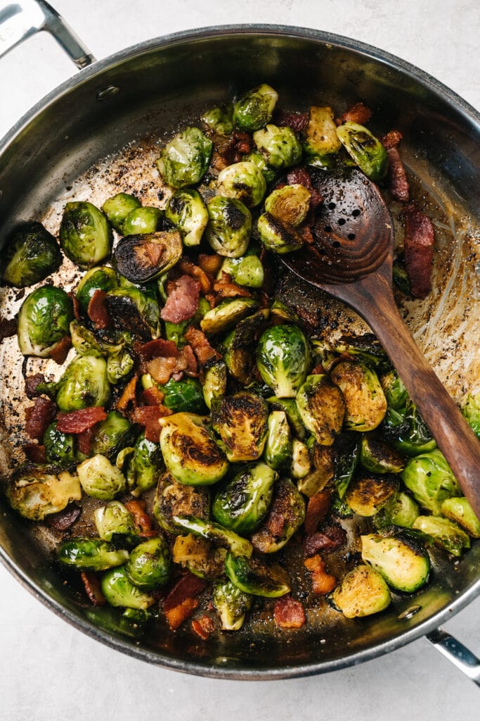 Crispy sautéed Brussels sprouts with bacon in a skillet with a wood spoon.