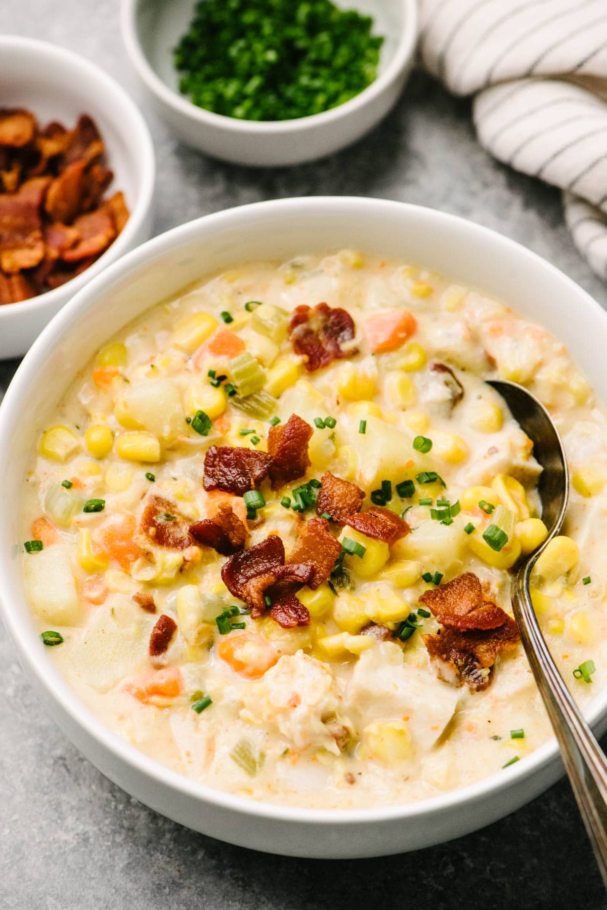 Side view, a spoon tucked into a bowl of turkey corn chowder on a concrete background; small bowls of bacon and chives are in the background, as well as a striped linen napkin.