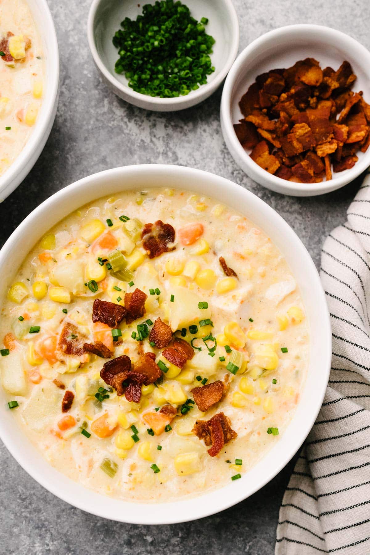 A bowl of turkey corn chowder garnished with bacon and chives on a concrete background; the bowl is surrounded by small bowls of cooked bacon and fresh chives, as well as a striped linen napkin.