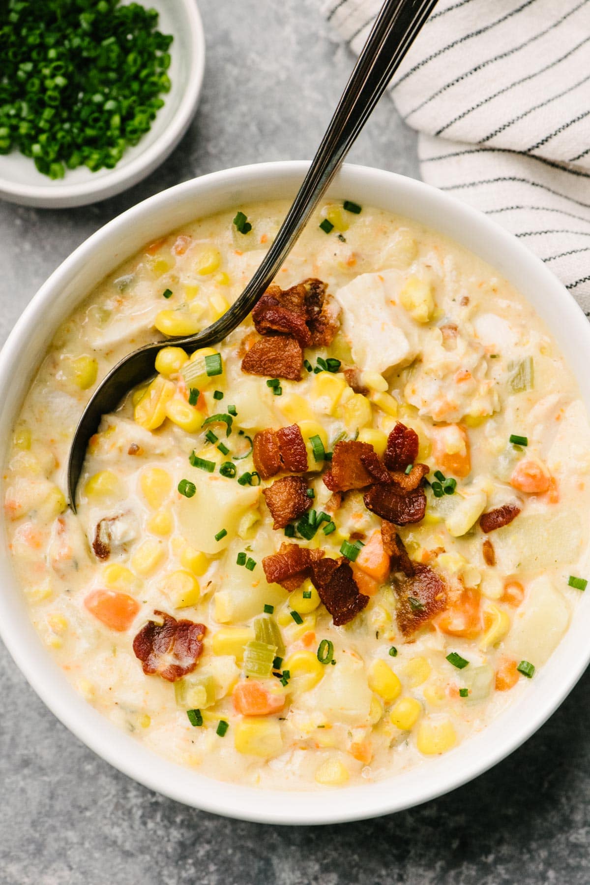 A soup spoon tucked into a bowl of turkey corn chowder with a bowl of chives and striped linen napkin tucked to the side.