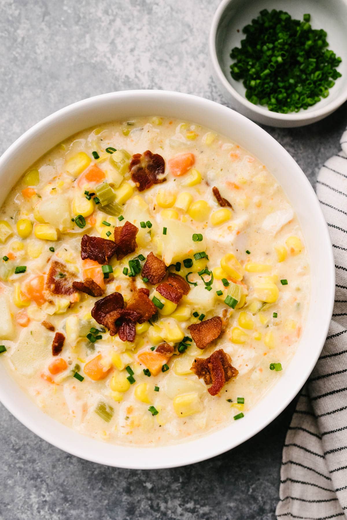 A bowl of turkey corn chowder garnished with crispy bacon and fresh chives on a concrete background; a striped linen napkin and small bowl of fresh chives are to the side.