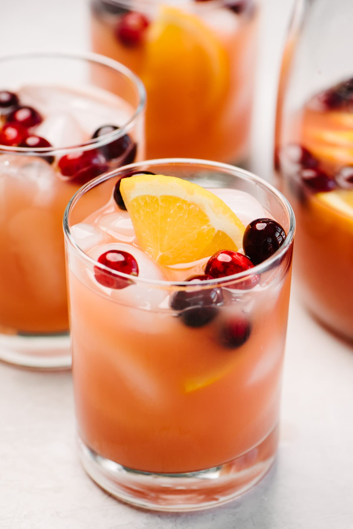 Side view, three glasses of whiskey punch next to a glass pitcher on a cement background; each cocktail is garnished with fresh cranberries and orange slices.