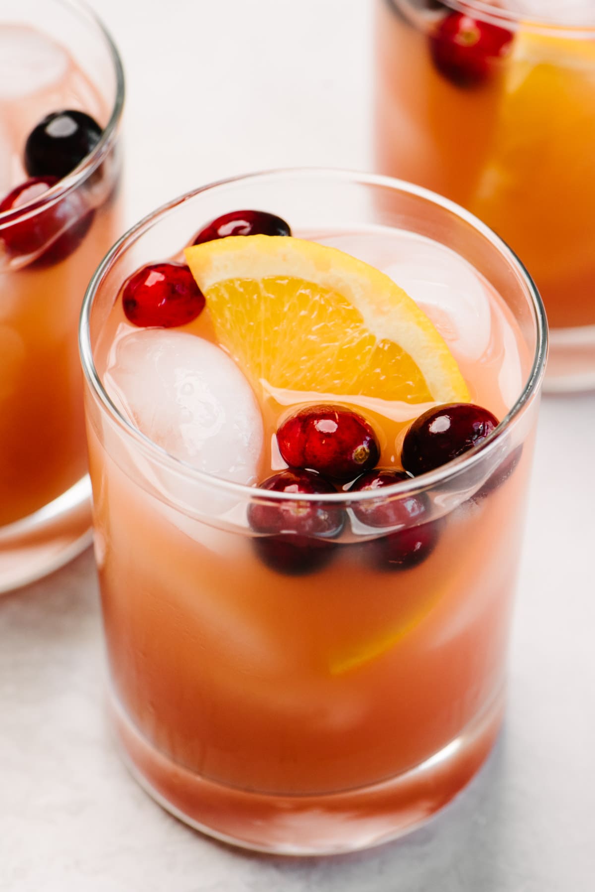 Side view, a glass of whiskey punch made with cranberry and orange juice; another cocktail and a glass pitcher are in the background.