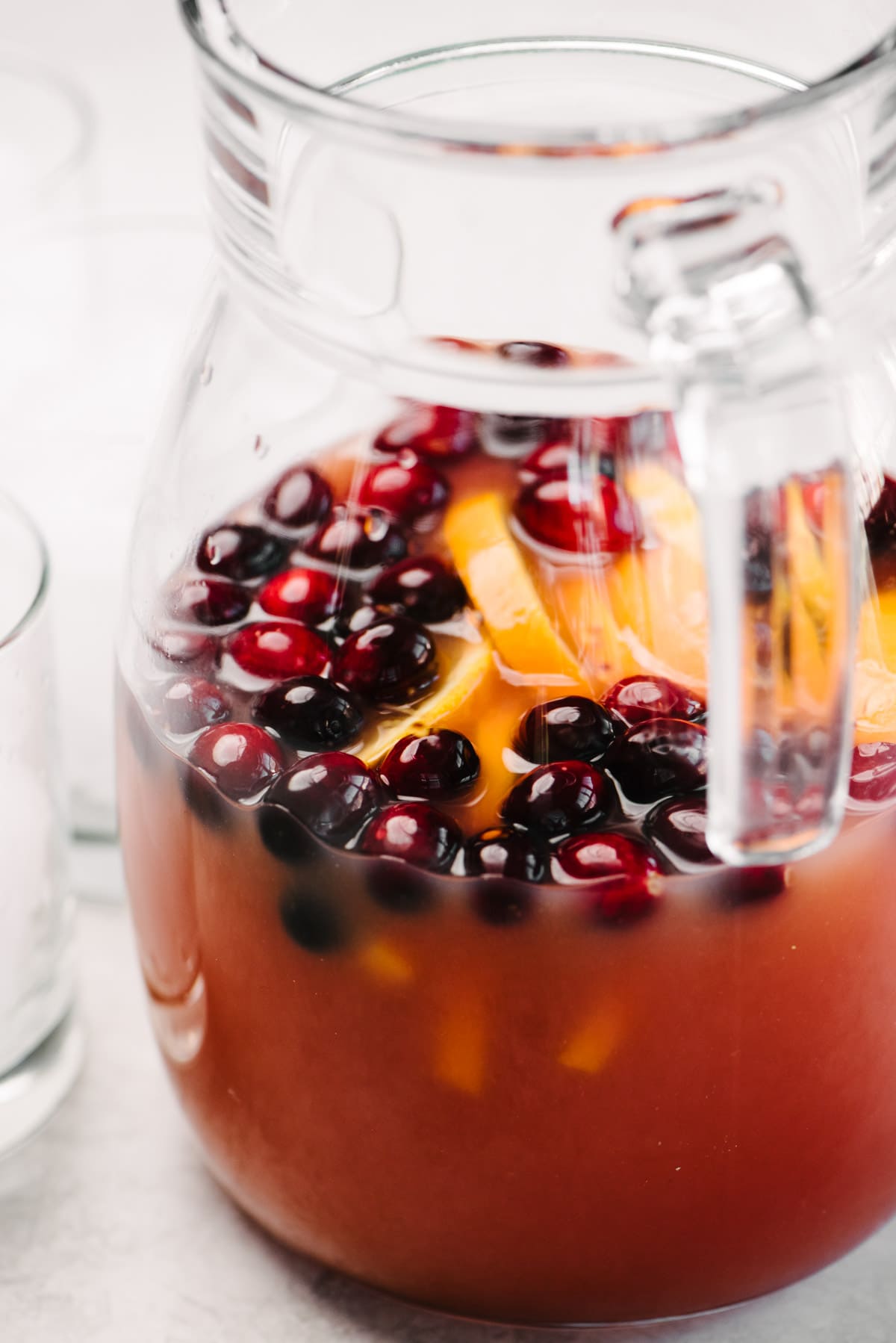 Side view, a sour whiskey punch in a large glass pitcher garnished with orange slices and fresh cranberries.
