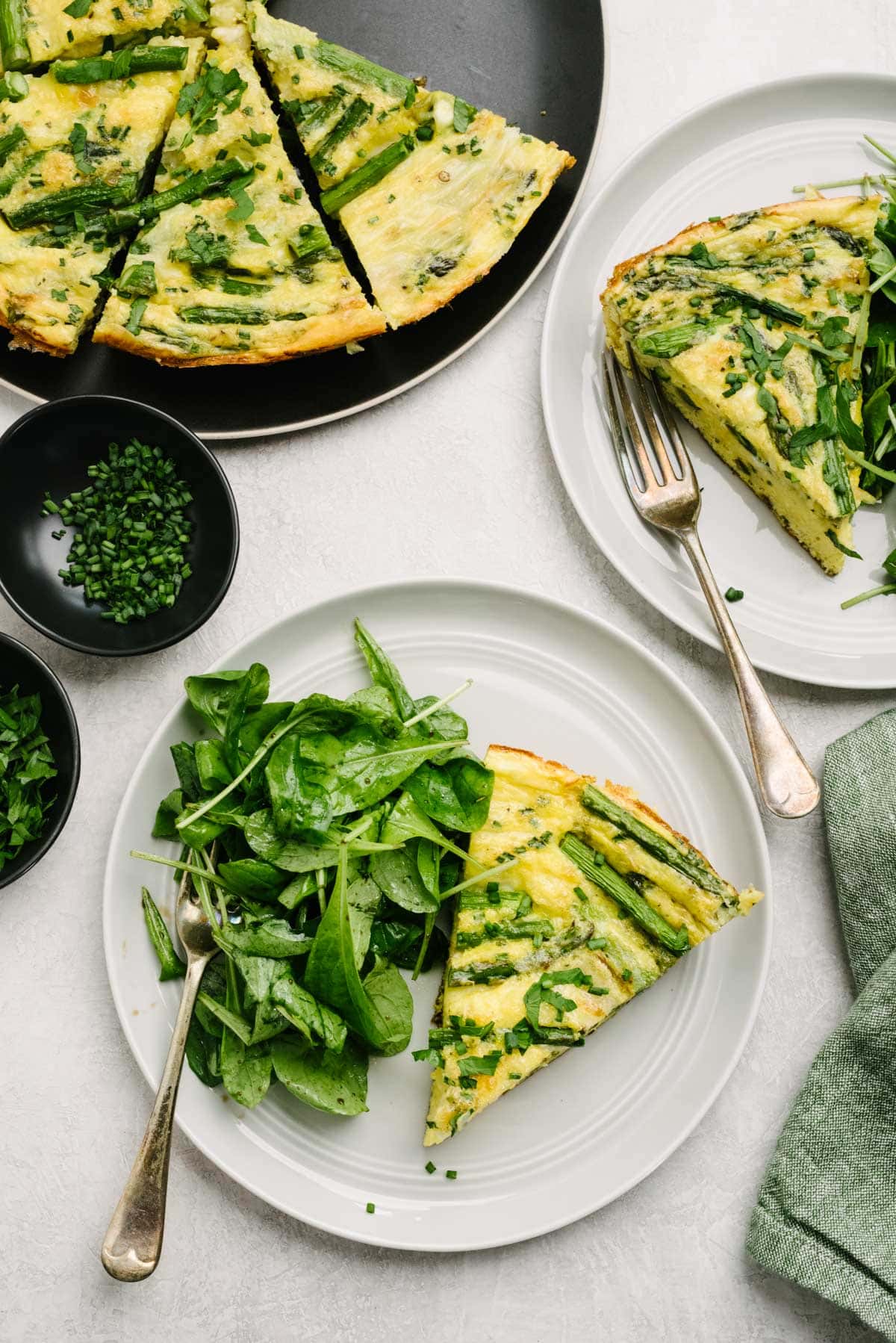 Two plates with asparagus frittata slices and tossed green salads as well as a cast iron skillet with the remaining frittata slices on a concrete background; a green linen napkin and small bowls filled with chopped fresh chives and parsley surround the plates and skillet.