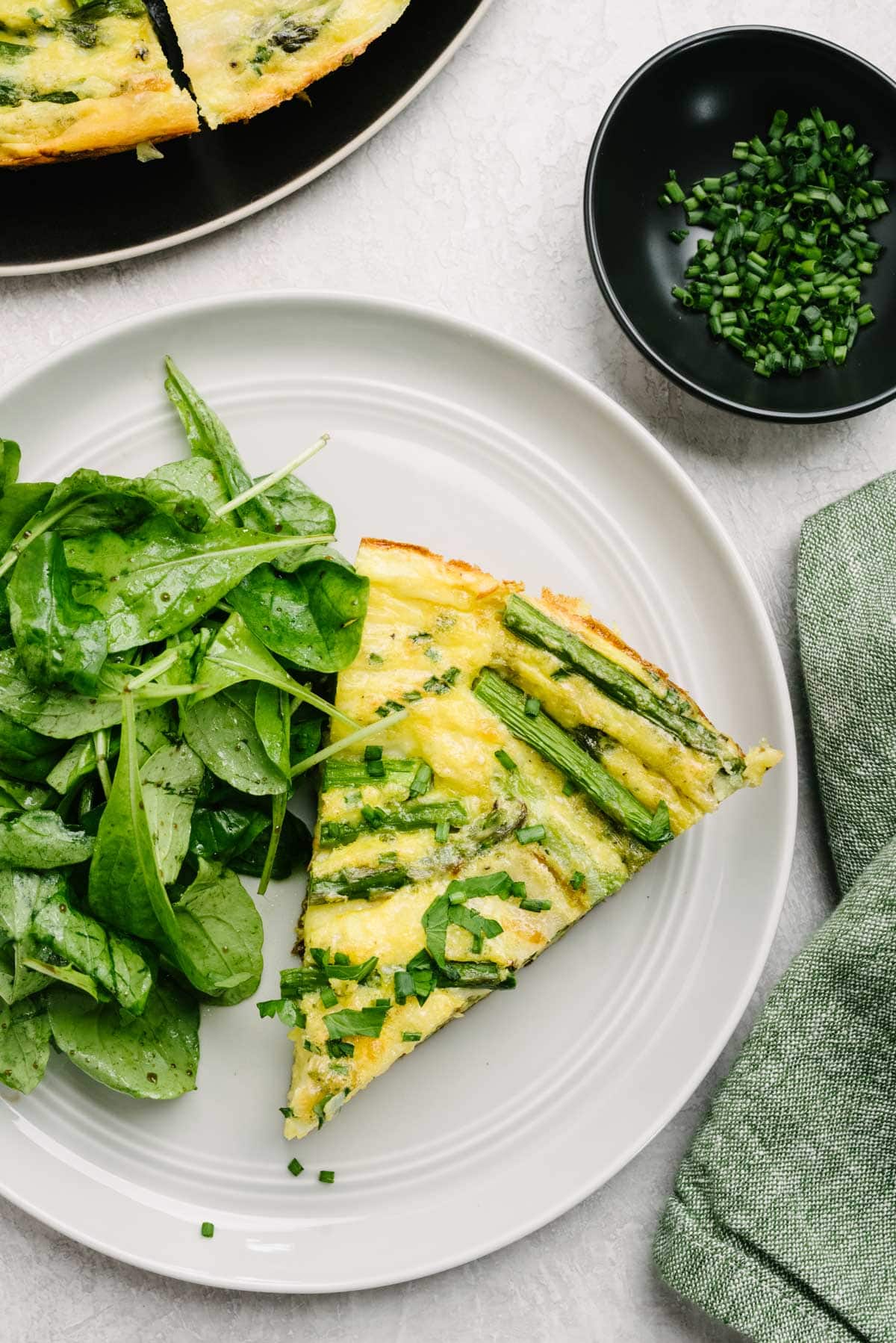 A slice of asparagus frittata on a grey plate with a tossed green salad; a bowl of chopped parsley and a green linen napkin surround the plate.