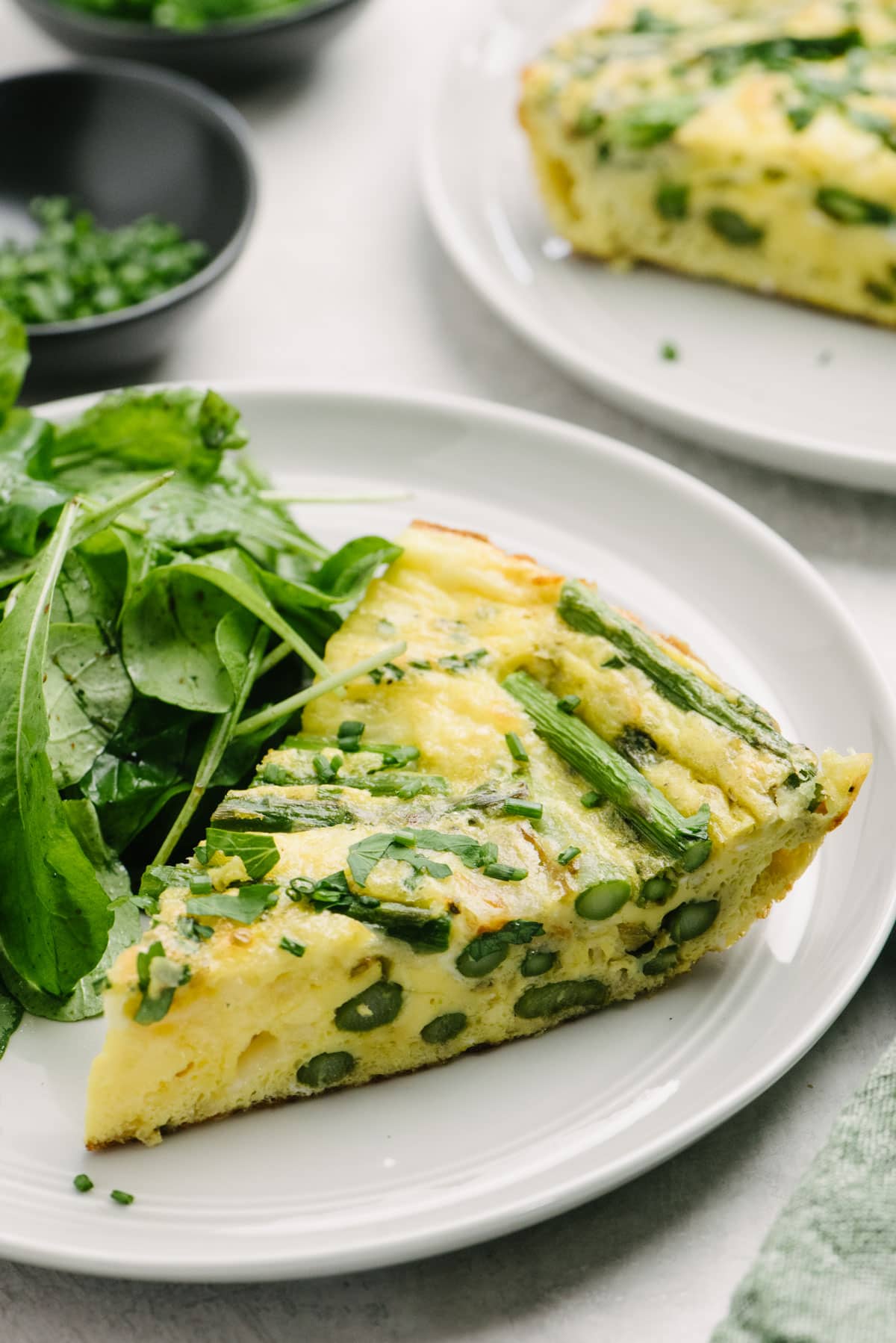 Side view, a slice of asparagus frittata on a light grey plate with a tossed green salad; another plate of frittata as well as a small bowl of chopped chives are in the background.