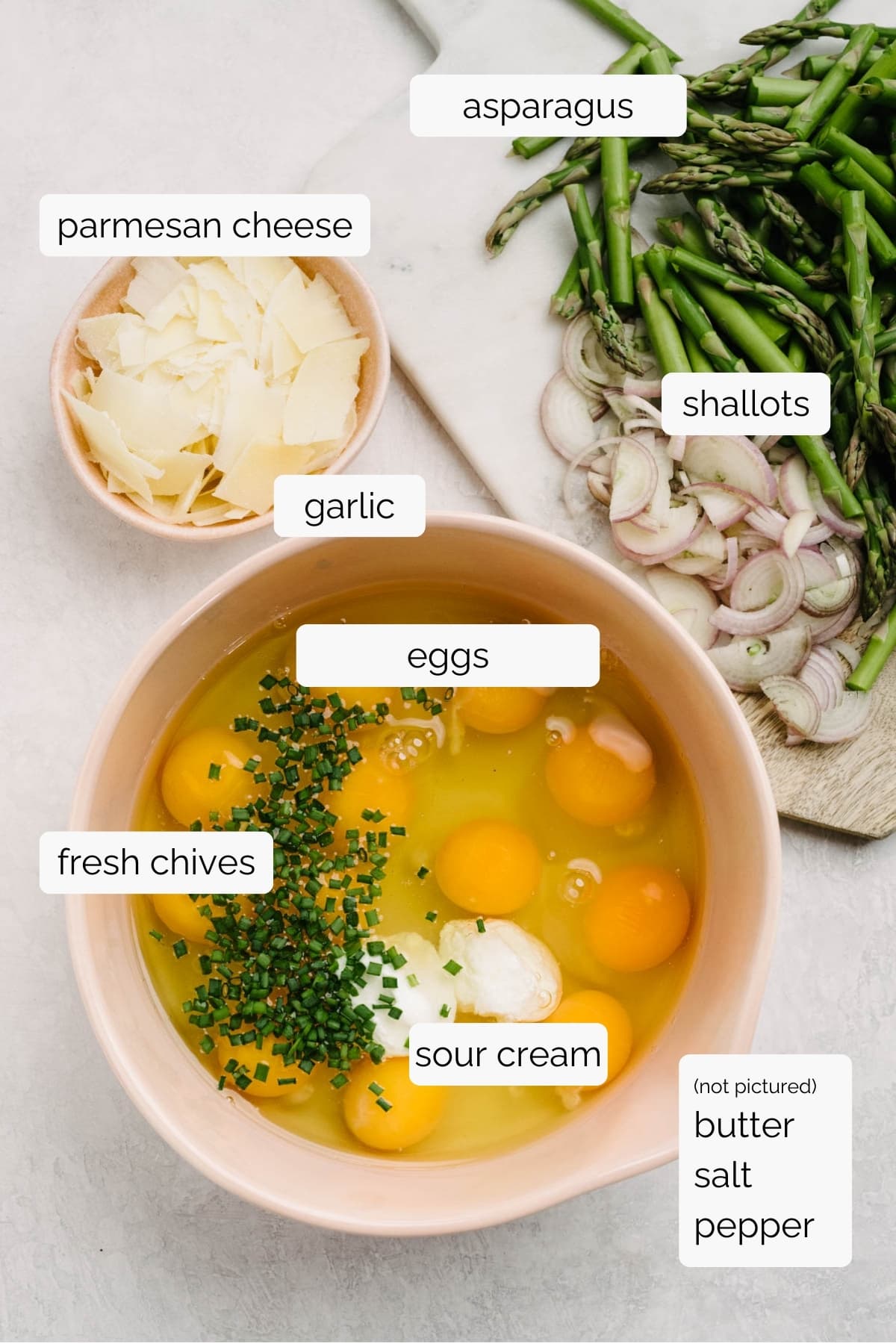 The ingredients for an asparagus frittata arranged on a concrete background - eggs, fresh chives, and sour cream in a large mixing bowl; shaved parmesan cheese in a small bowl; chopped asparagus and thinly sliced shallots on a cutting board.