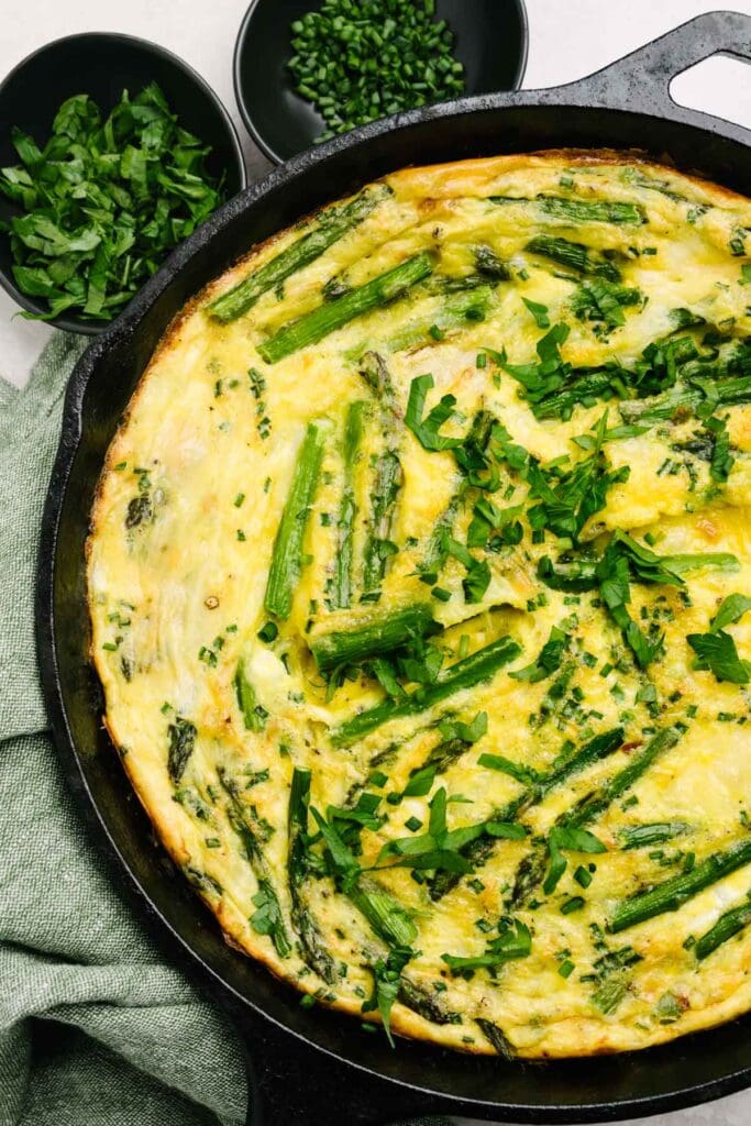 As asparagus frittata in a cast iron skillet on a concrete background, surrounded with a green linen napkin and small pinch bowls of fresh chopped parsley and chives.