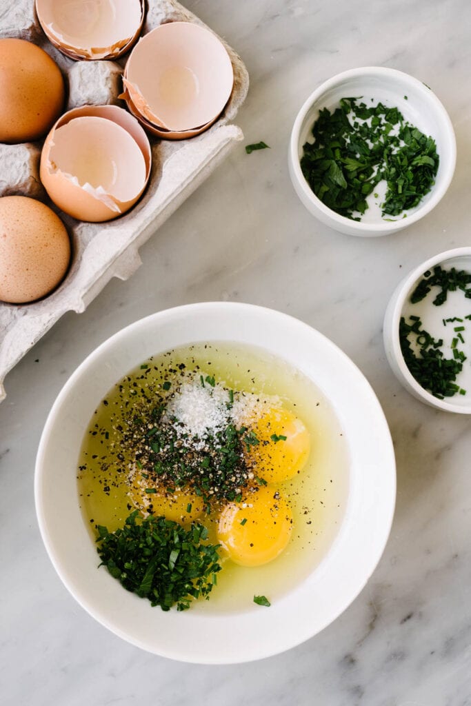 Whole, fresh parsley, fresh chives, salt, and pepper in a small white bowl on a marble background surrounded by a carton of eggs and small white dishes filled with chopped herbs.