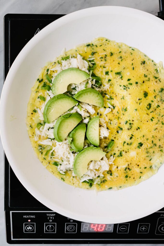 The interior of an omelet in a skillet over a burner; one half is layered with parmesan cheese, crab meat, green onions, and sliced avocado.