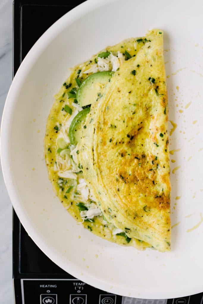 An American-style crab omelet in a skillet - one half is folded over the filling, which includes crab meat, parmesan cheese, avocado, and green onions.