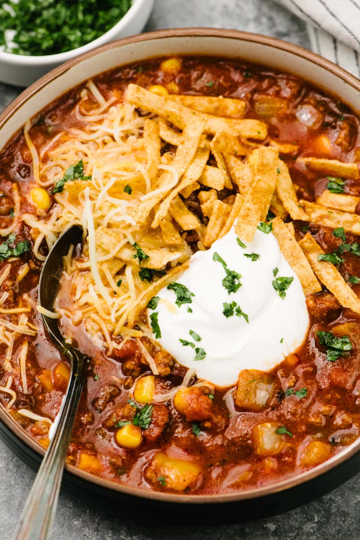 A soup spoon tucked into a bowl of crockpot taco soup, garnished with sour cream, shredded cheese, tortilla strips, and chopped cilantro; a small bowl of chopped cilantro and a striped linen napkin are in the background.
