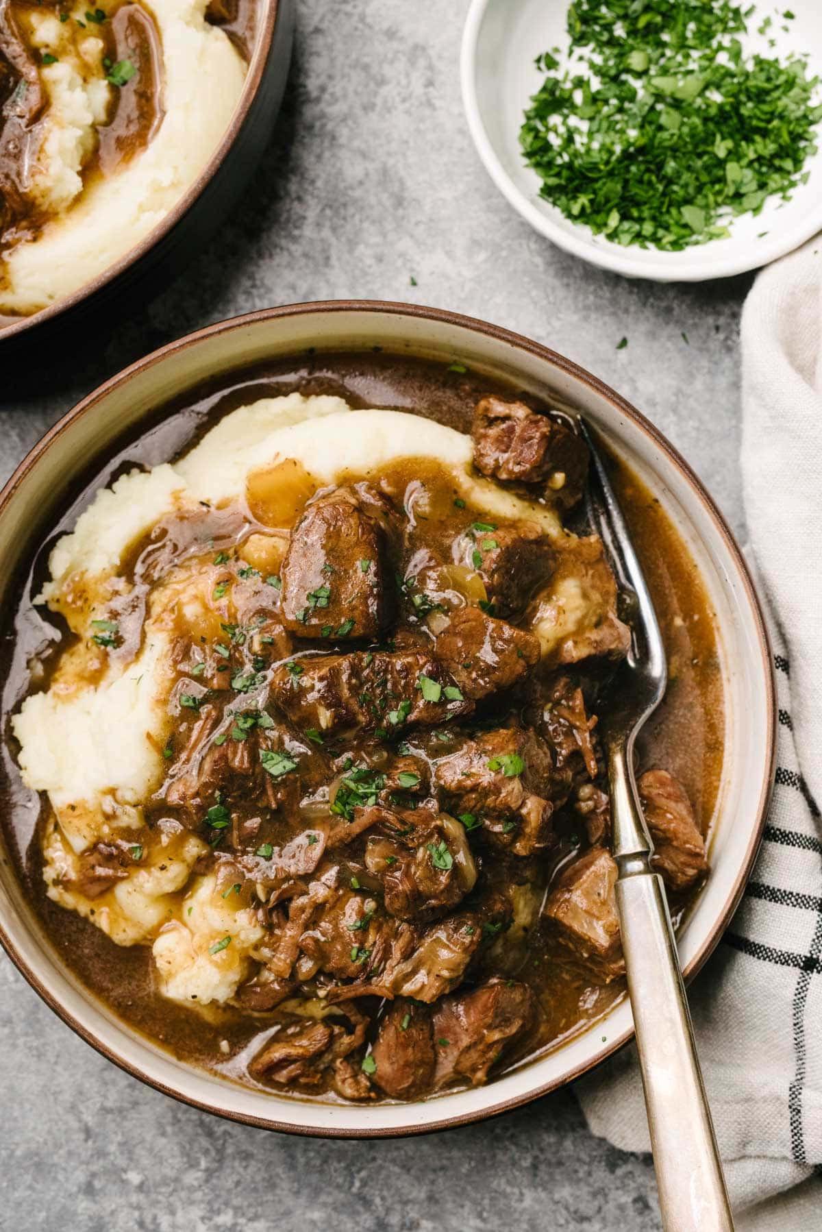 A fork tucked into Instant pot beef tips over mashed potatoes in a low serving bowl on a concrete background, surrounded by a striped linen napkin and small bowl of finely chopped parsley.