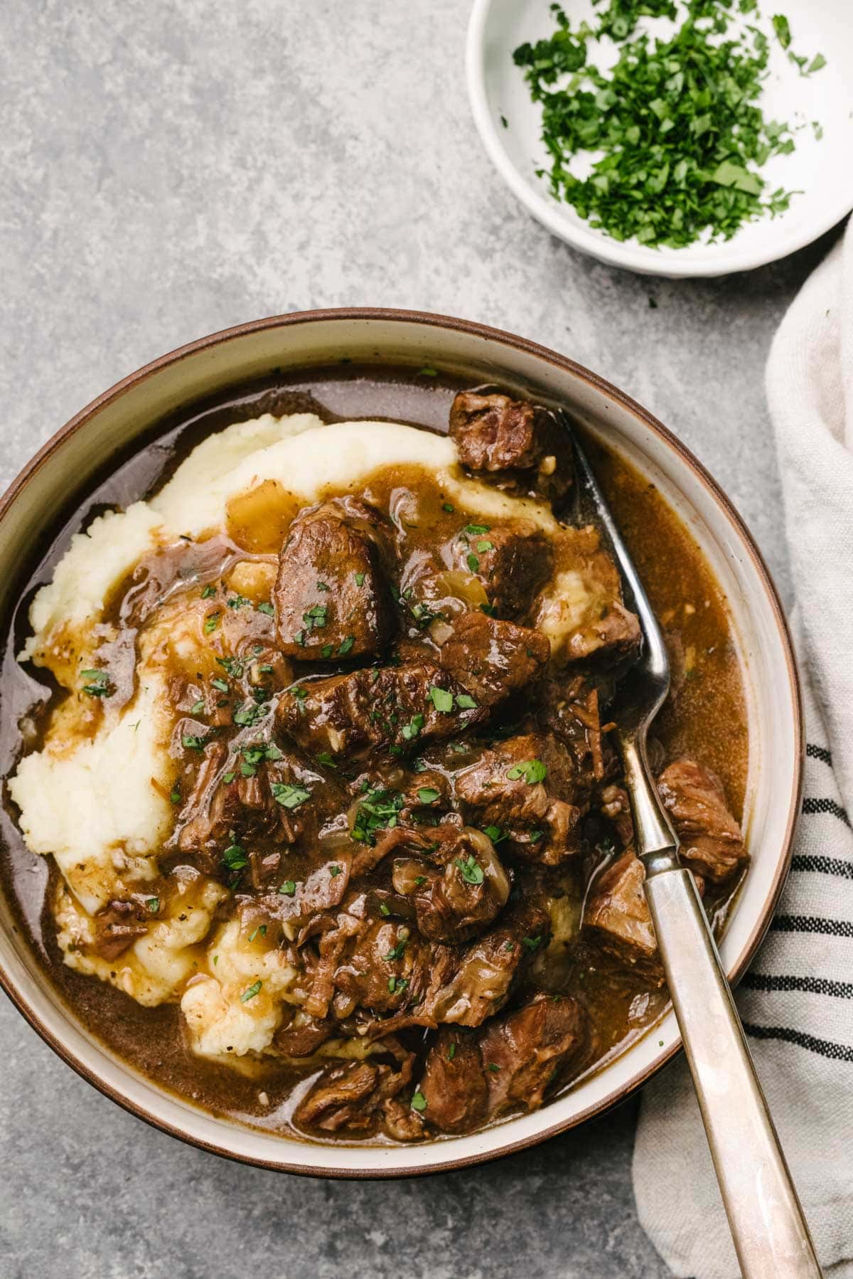 A fork tucked into tender Instant Pot beef tips over mashed potatoes in a low shallow bowl; the bowl is surrounded by a striped linen napkin and small bowl of chopped fresh parsley.