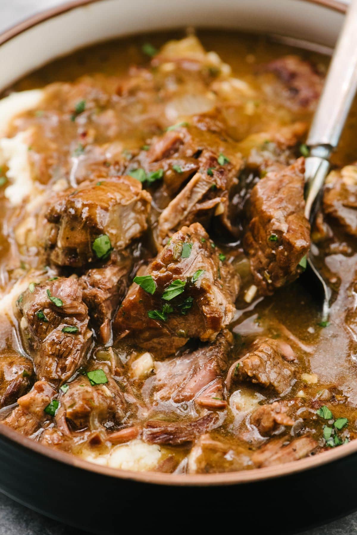 Side view, a fork tucked into fall-apart-tender beef tips over mashed potatoes in a wide, low serving bowl.