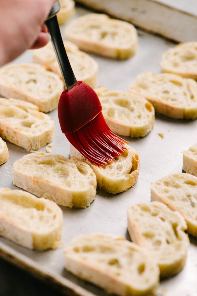 Side view, brushing olive oil onto thinly sliced pieces of bread arranged on a baking sheet.