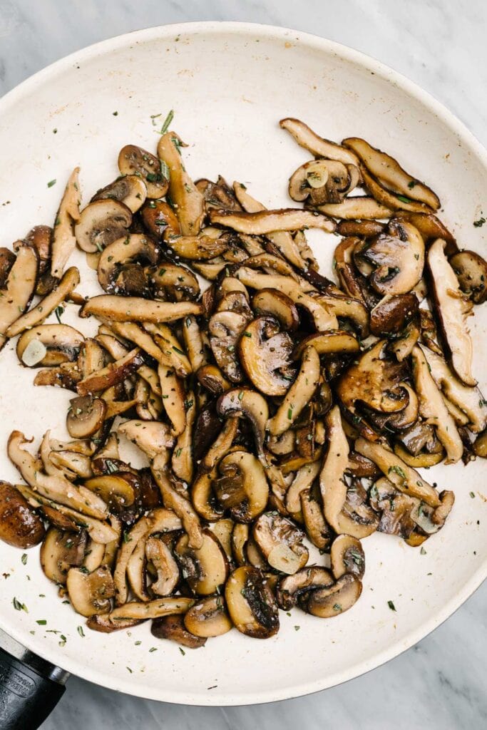 Sautéed mushrooms in a skillet seasoned with thinly sliced garlic and fresh rosemary.