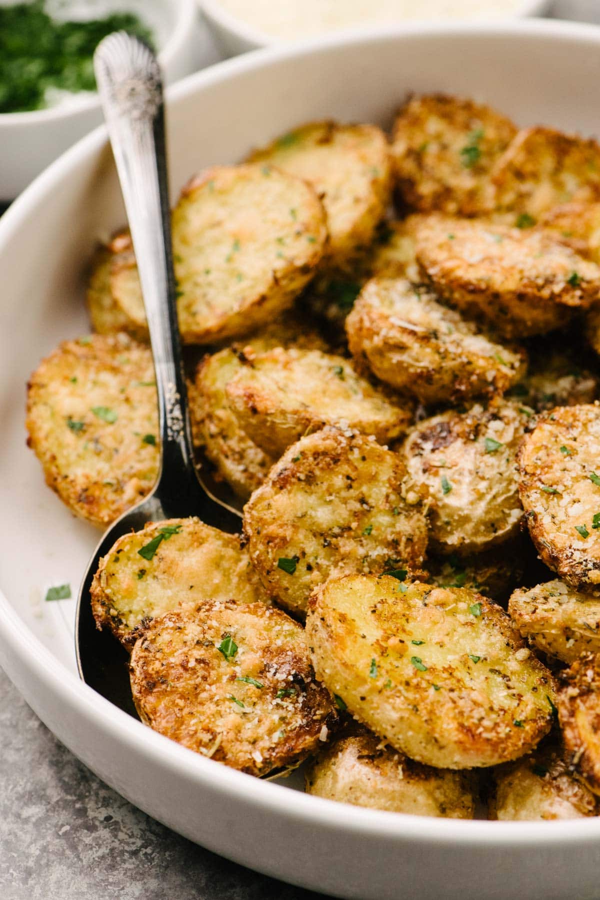 Side view, a silver serving spoon tucked into a white serving bowl of crispy roasted parmesan potatoes; small dishes of grated parmesan cheese and finely chopped parsley are in the background.