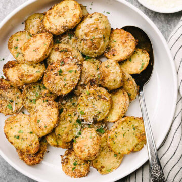 A silver serving spoon tucked into a bowl of crispy parmesan crusted potatoes, garnished with fresh chopped parsley.