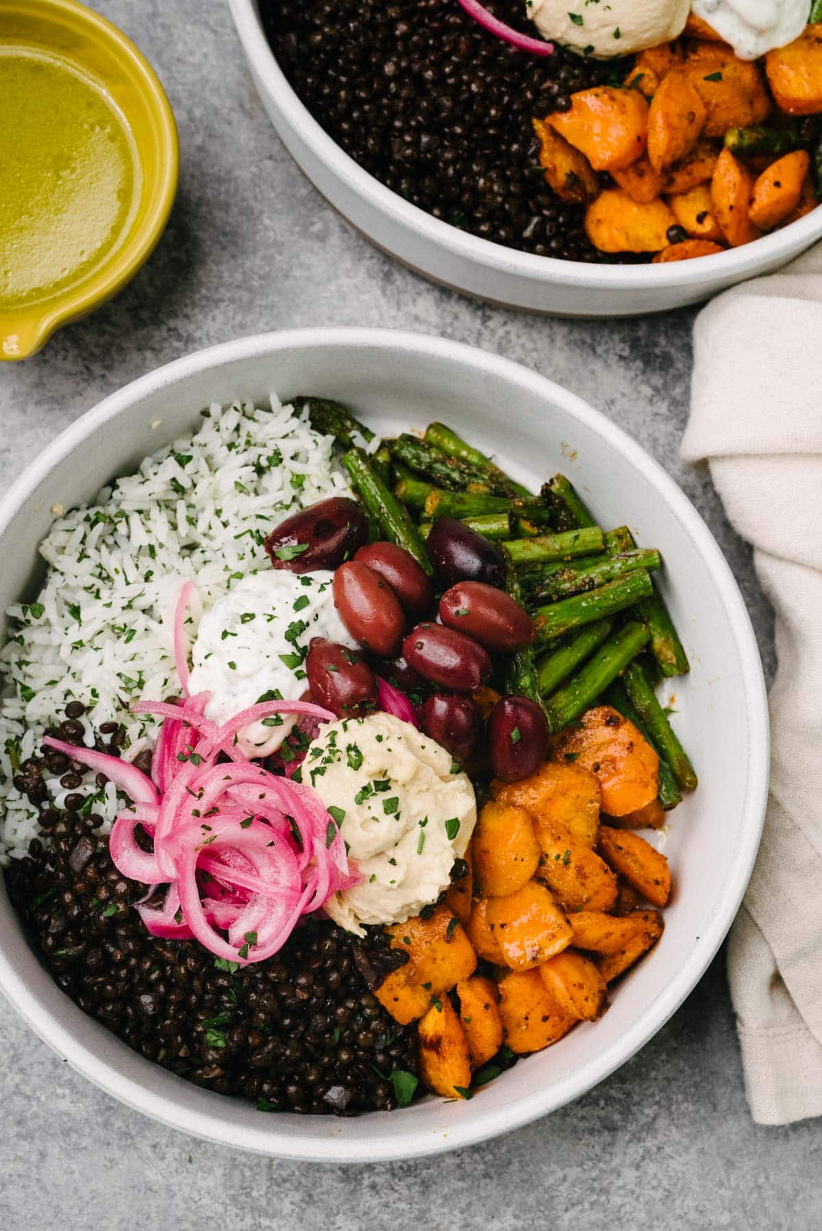 Two black lentil bowls on a concrete surface, surrounded by a cream linen napkin and yellow spouted bowl filled with lemon dressing; each bowl is made with black lentils, herbed rice, roasted vegetables, and various toppings.