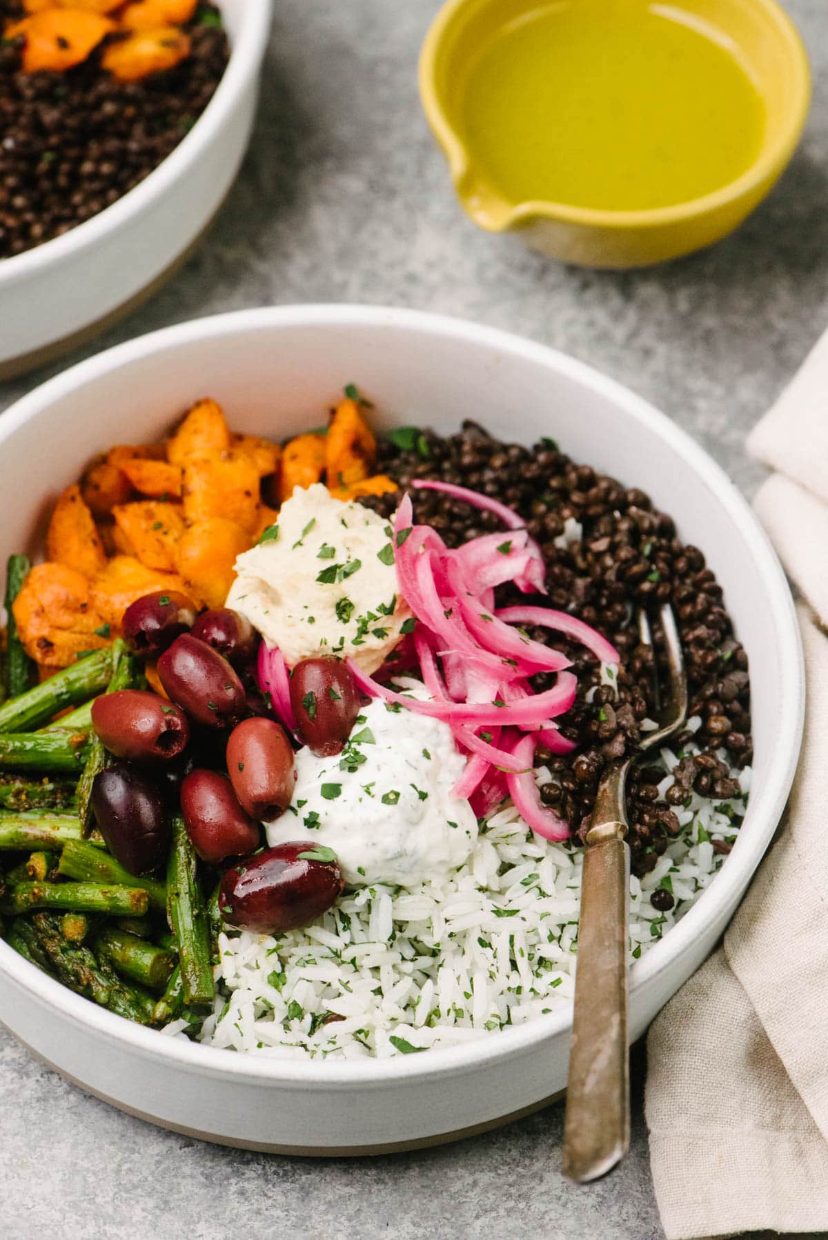 Side view, a fork tucked into a low serving bowl filled with black lentils, herbed rice, and roasted vegetables topped with olives, pickled red onions, and hummus; a cream linen napkin and small yellow bowl with lemon vinaigrette surround the bowl.