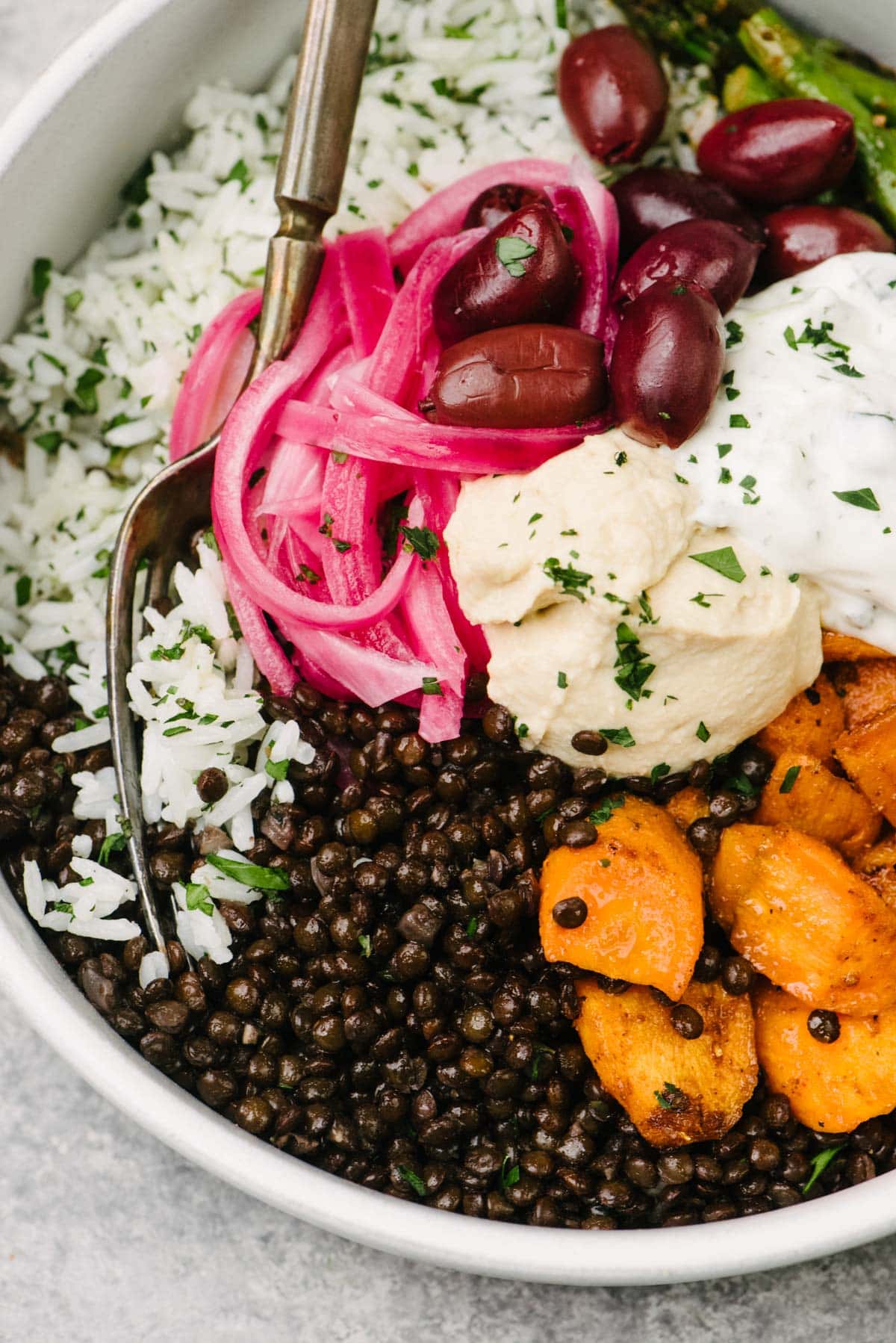 A fork tucked into a CAVA inspired mediterranean bowl made with black lentils, herb rice, and roasted vegetables topped with pickled red onions, kalamata olives, hummus, and tzatziki sauce.