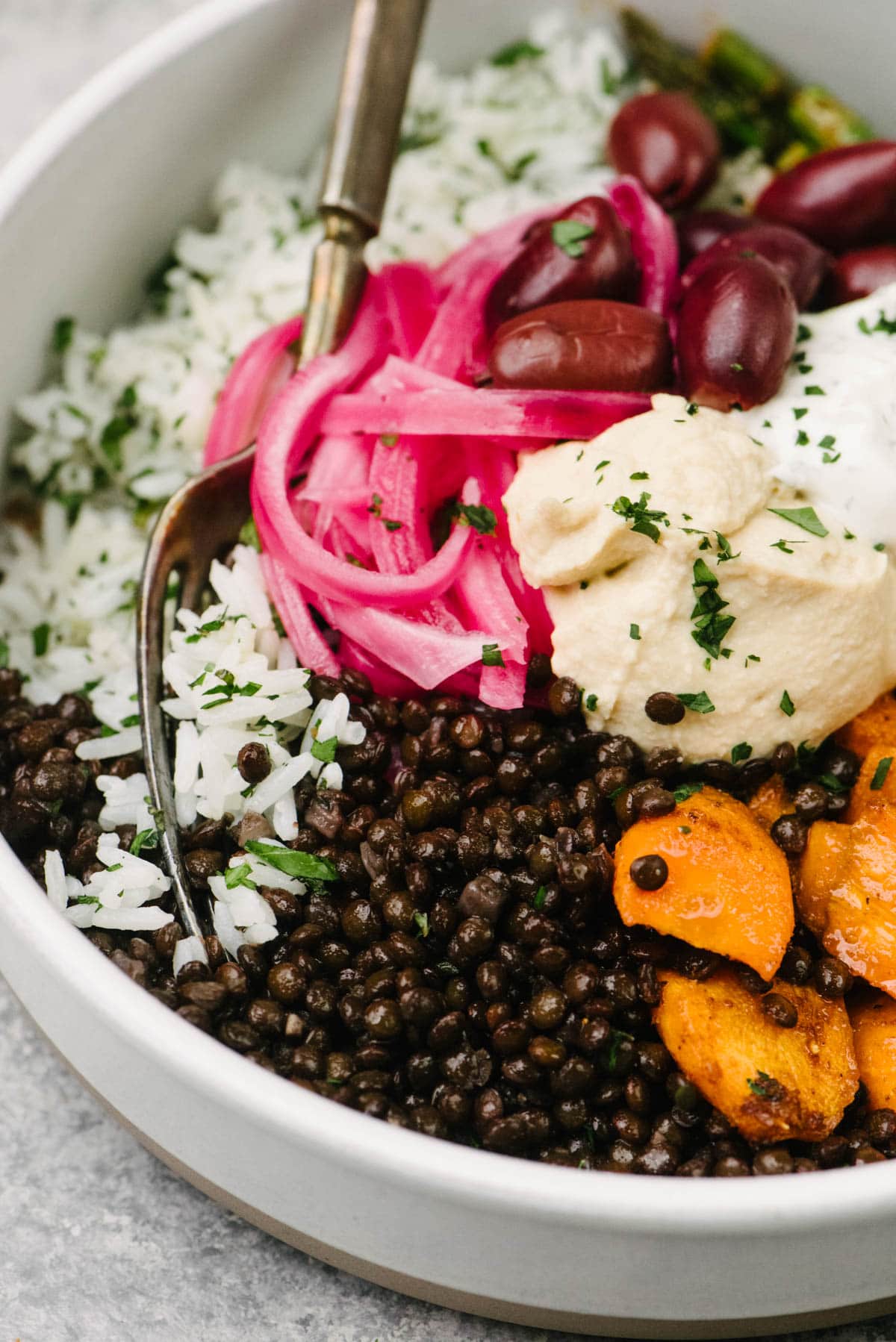 Side view, a fork tucked into a CAVA style black lentil bowl made with herbed white rice, black lentils, and roasted carrots and asparagus topped with hummus, olives, and pickled red onions.