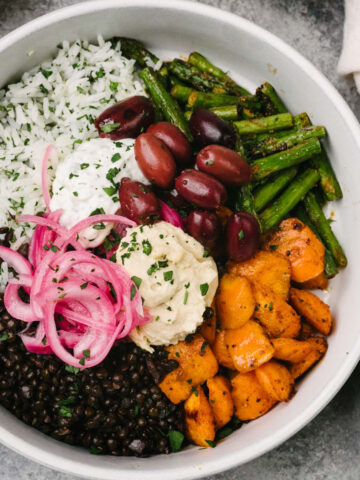 A mediterranean grain bowl made with black lentils, herb rice, and roasted vegetables, topped with hummus, tzatziki, kalamata olives, and pickled red onions; a cream linen napkin is tucked to the side.