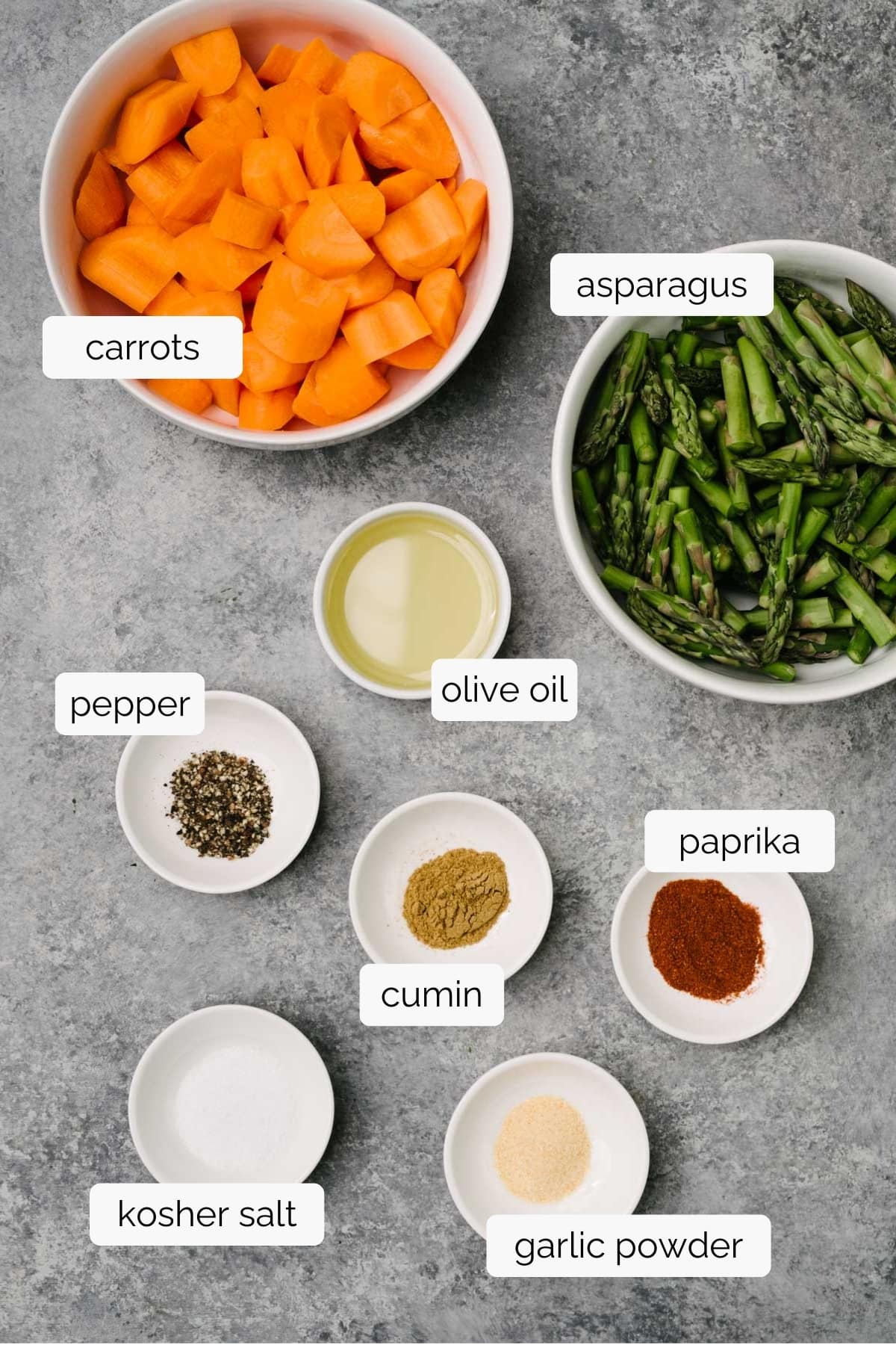 The ingredients for roasted carrots and asparagus in small bowls arranged on a concrete background - sliced carrots, chopped asparagus, olive oil, salt, pepper, paprika, cumin, and garlic powder.