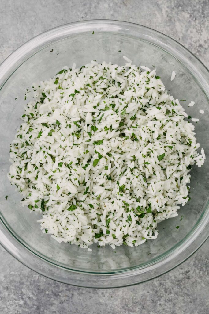 Cooked white rice tossed with finely chopped fresh mint and fresh parsley in a glass mixing bowl.